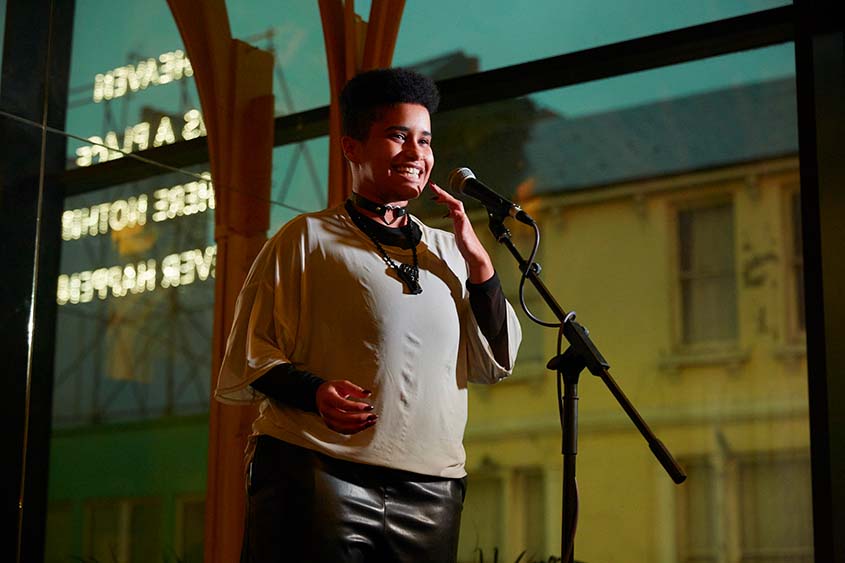 Photograph: A mid-shot of a person smiling and speaking into a microphone on stage. They are a Black person with short hair on top and shaved sides. They are wearing a black leather like skirt, a white t-shirt over a black top. A black collar with a metal heart in the centre of their throat and another black pendant necklace. Behind them is a large window. It is dark outside but there it a large sign with backwards writing lit up.